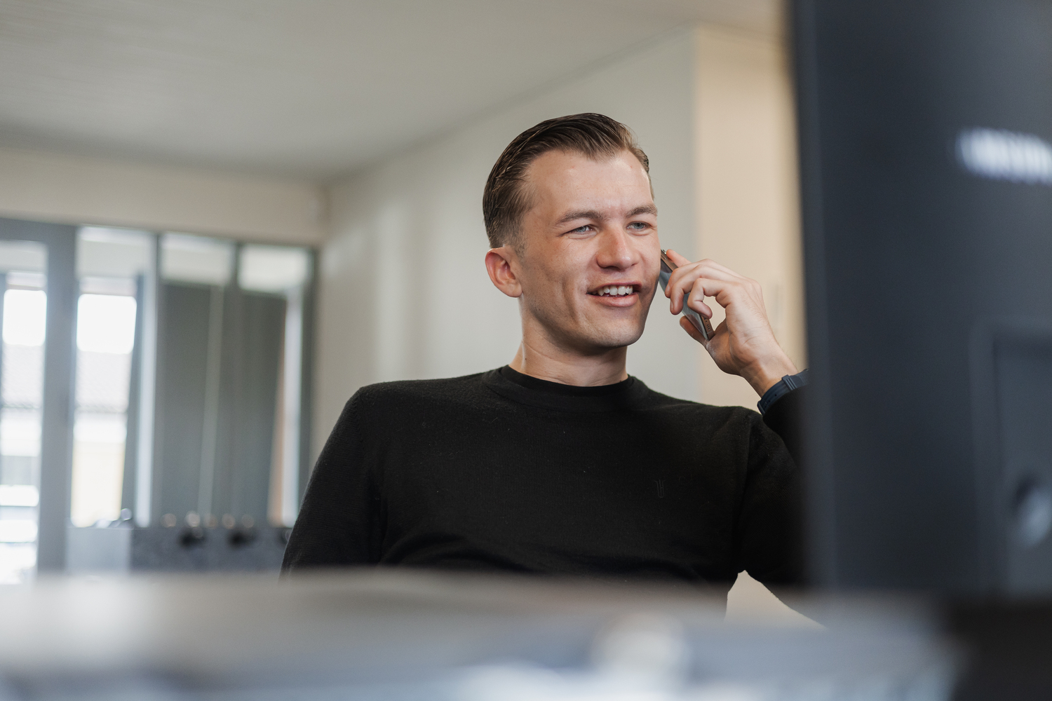 Mand i sort trøje taler i telefon på kontor