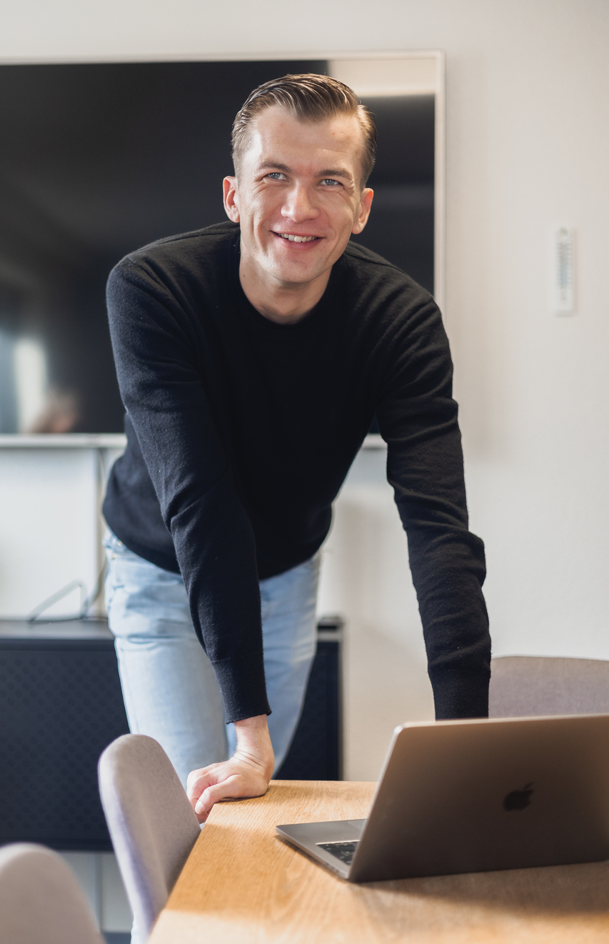 Mand i sort sweater læner sig over et bord med en bærbar computer, smilende og ser direkte ind i kameraet.